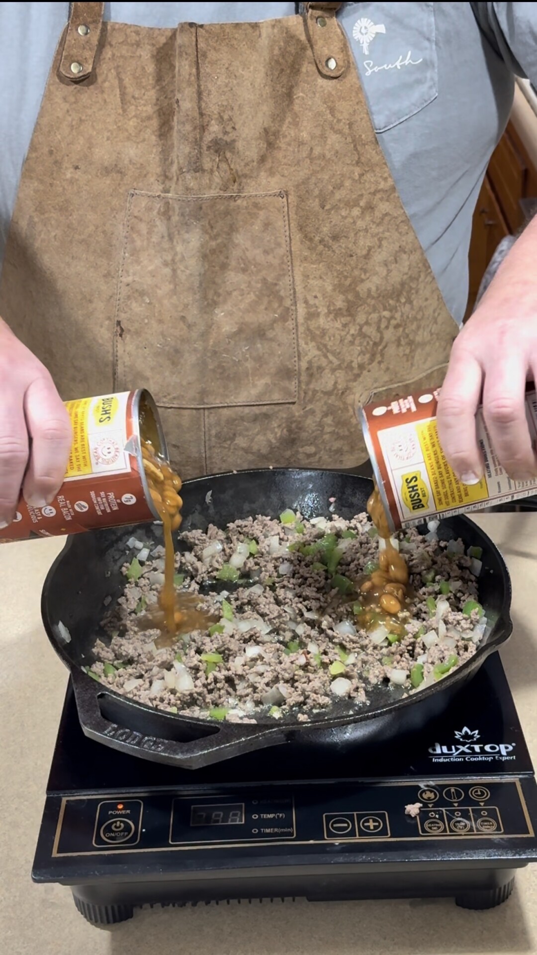 A person in a brown apron pours canned beans into a cast iron skillet with cooked ground meat and chopped green bell peppers on an electric stovetop.