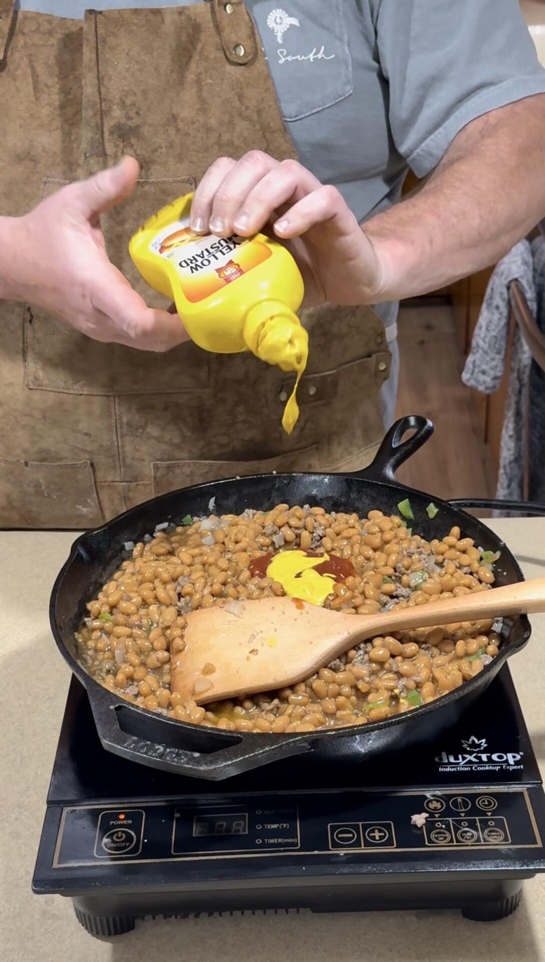 Person squeezing French’s yellow mustard onto baked beans in a cast iron skillet on an induction cooktop, with a wooden spatula resting in the beans. Person wears a brown apron and gray shirt.