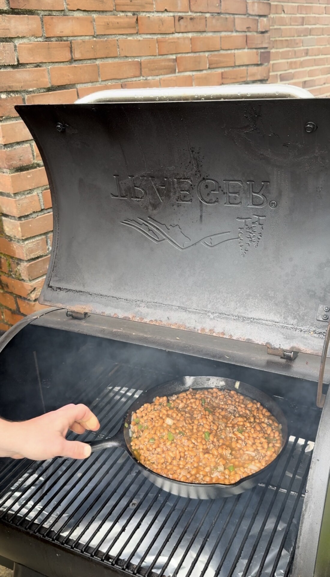 A hand stirs baked beans in a cast iron skillet on a smoker grill with the lid open; smoke is visible, and the grill has LONE STAR embossed on the inside of the lid. Brick wall in the background.