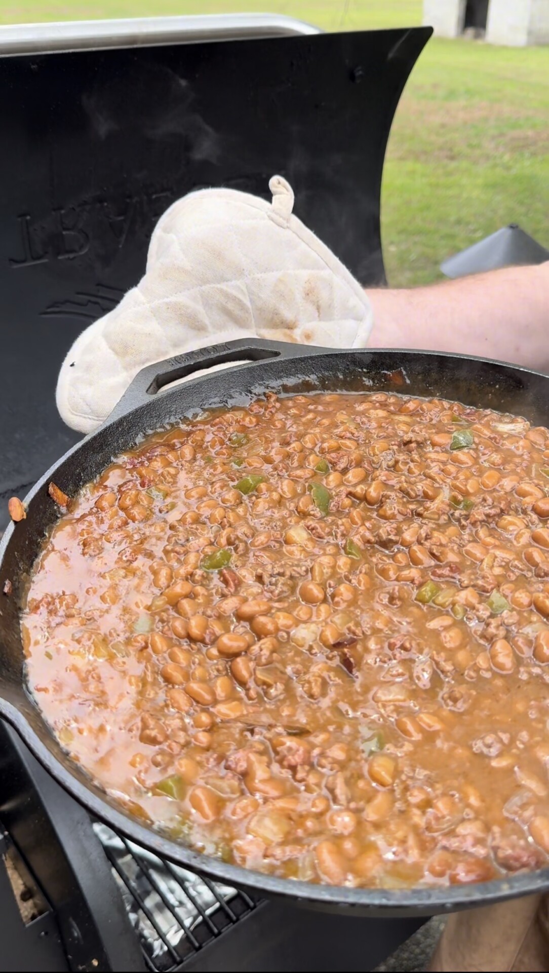 A close-up of baked beans simmering in a cast iron skillet on a grill, with an oven mitt holding the pan handle and green grass visible in the background.