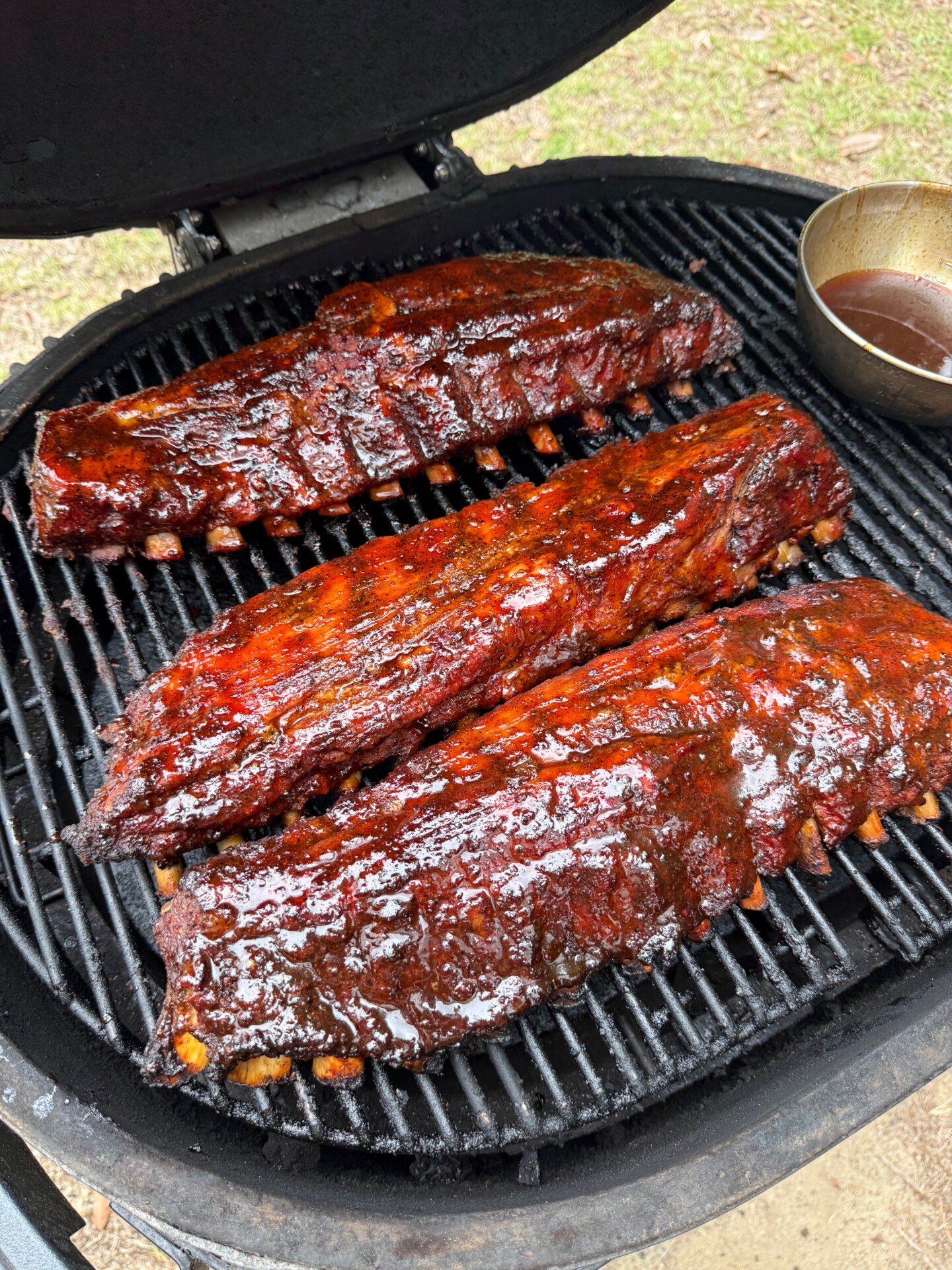 Three racks of glazed, barbecued ribs cooking on a round grill with grill marks visible. A small metal bowl with sauce sits in the upper right corner of the grill.