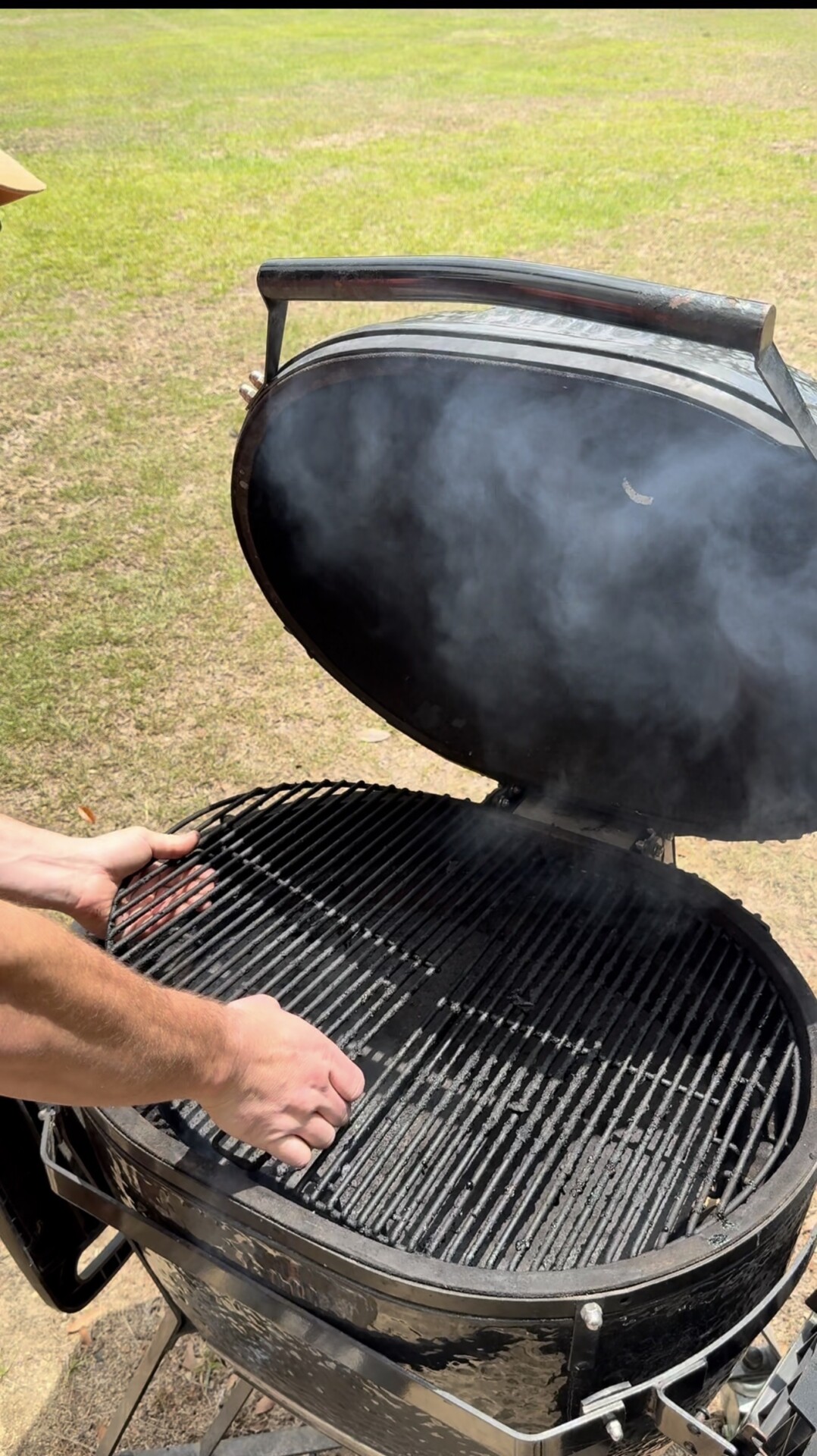 A person is placing the grill grate onto a black round barbecue grill that is emitting smoke, outdoors on a grassy area. The grill lid is open, and the scene suggests preparations for cooking.