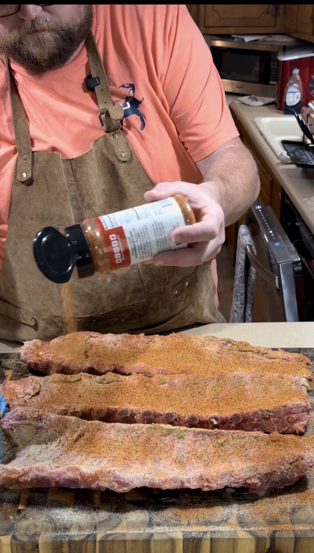 A person wearing a brown apron and pink shirt is sprinkling seasoning from a bottle onto slabs of raw ribs on a kitchen counter.