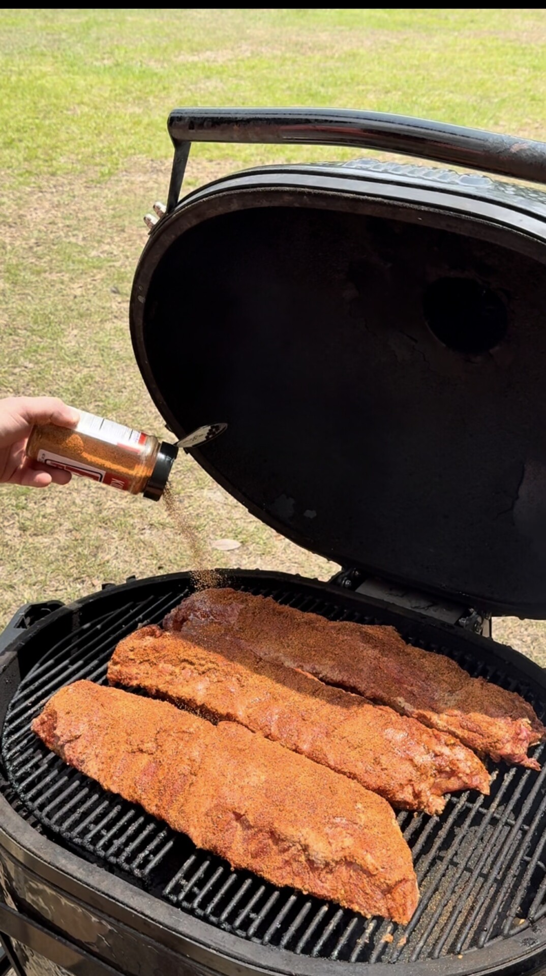 A hand is sprinkling seasoning onto three racks of ribs cooking on a grill with the lid open. The grill is outdoors on grass under daylight.