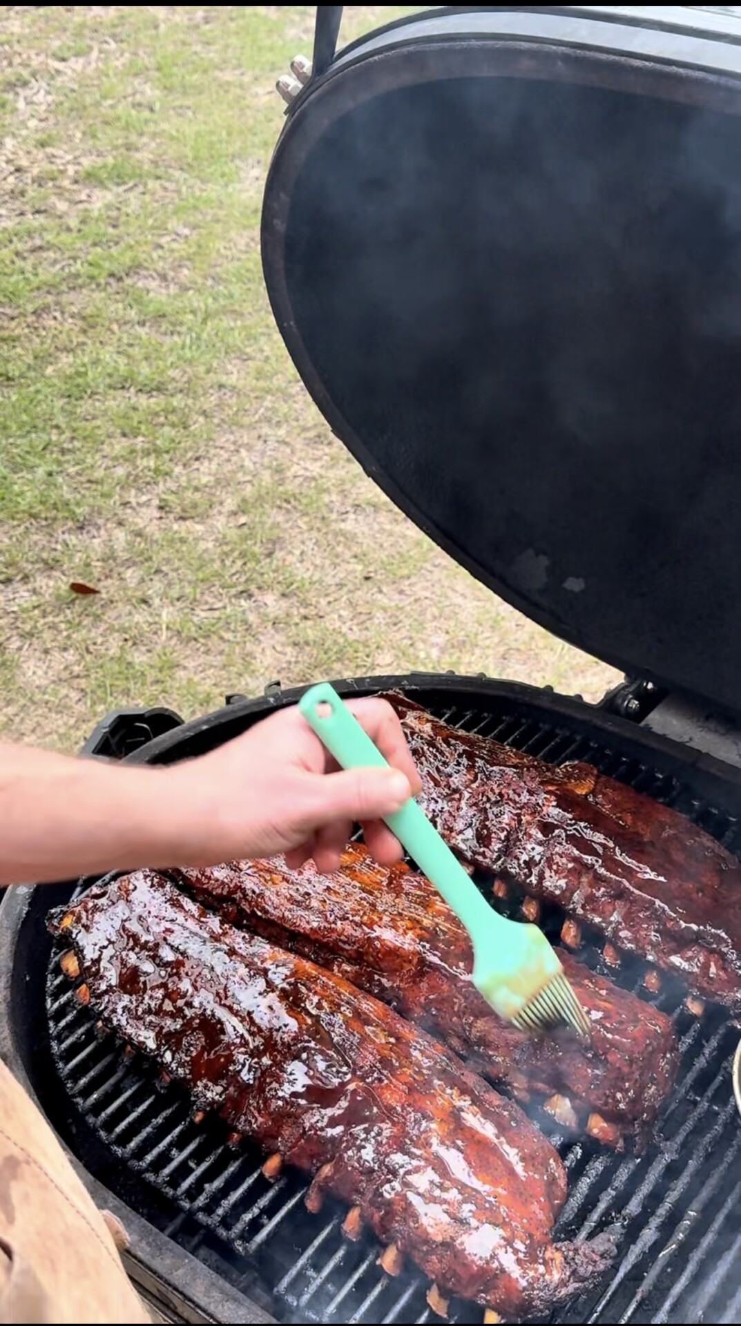 A person uses a green silicone brush to apply sauce to several racks of ribs cooking on a grill outdoors, with smoke rising and grass visible in the background.