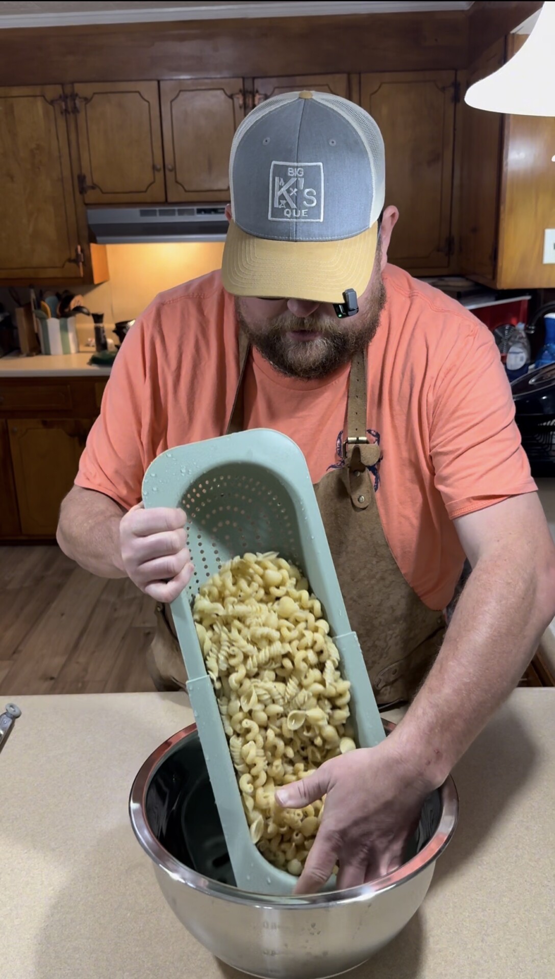 A man wearing an apron and a cap drains cooked pasta using a blue colander over a metal bowl in a kitchen with wooden cabinets.
