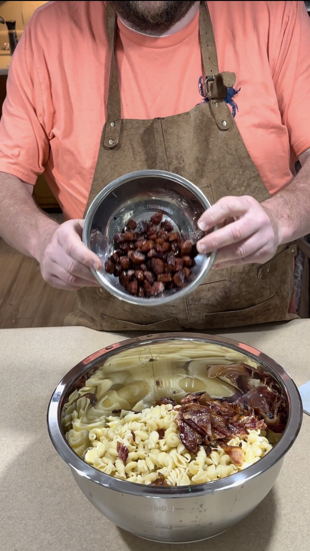 A person wearing a brown apron and orange shirt is holding a bowl of kidney beans above a larger bowl containing cooked pasta and bacon pieces on a kitchen counter.