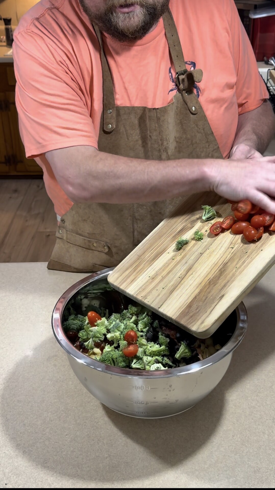A person in a brown apron and coral shirt is adding cherry tomatoes from a wooden cutting board into a large metal bowl filled with broccoli and vegetables on a kitchen counter.