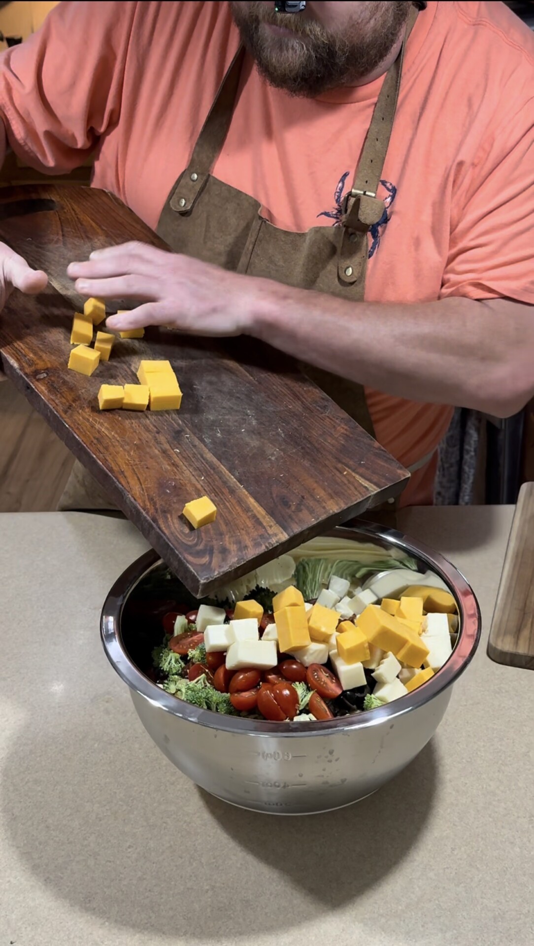 A person in an apron adds cubed cheddar cheese from a wooden board into a metal bowl filled with salad ingredients, including broccoli, grape tomatoes, and celery.