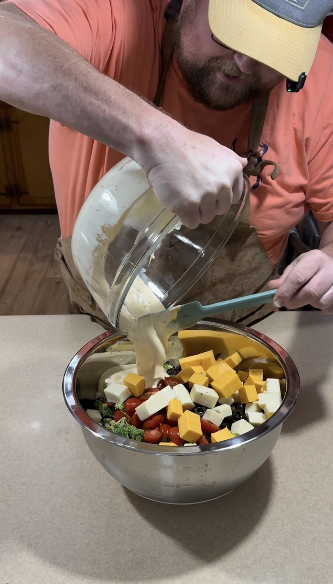 A person in an orange shirt pours creamy dressing from a glass bowl into a metal bowl filled with cubed cheese, cherry tomatoes, olives, and broccoli on a kitchen counter.