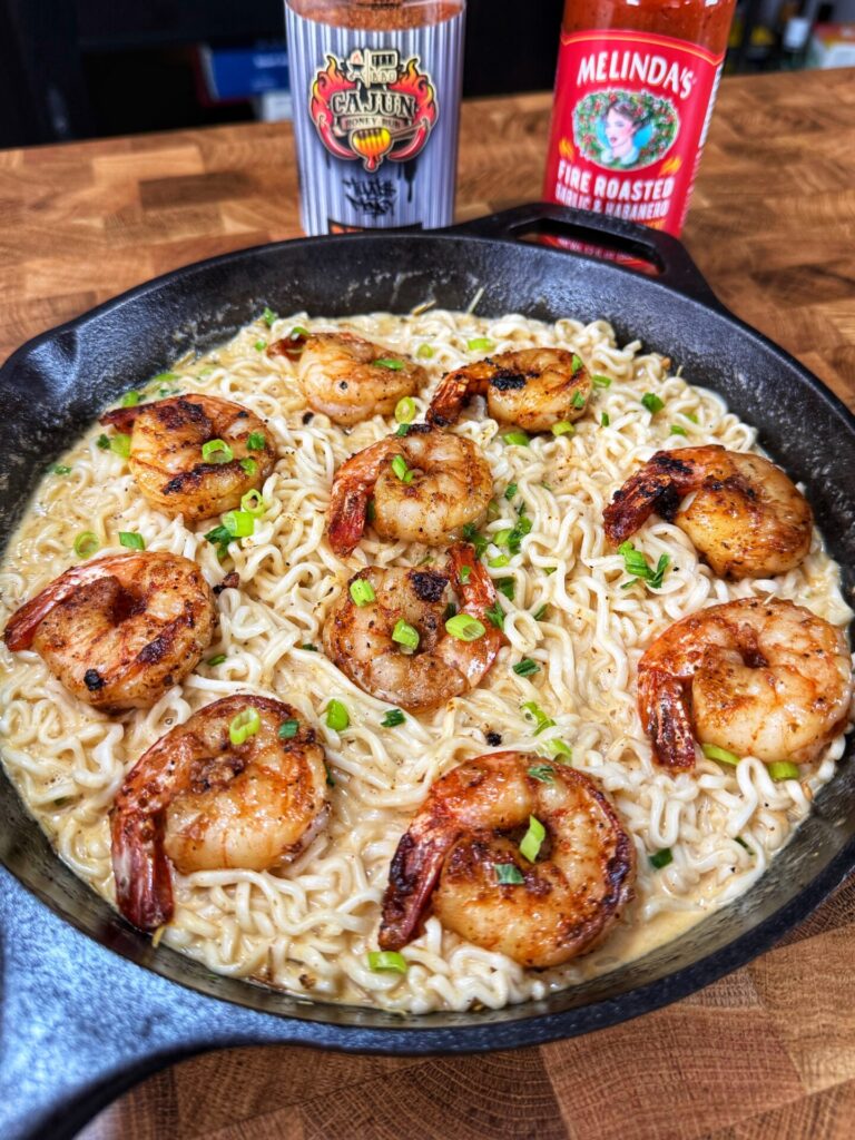 A skillet filled with cooked ramen noodles in a creamy sauce, topped with large seasoned shrimp and chopped green onions. Bottles of Cajun seasoning and hot sauce are visible in the background.