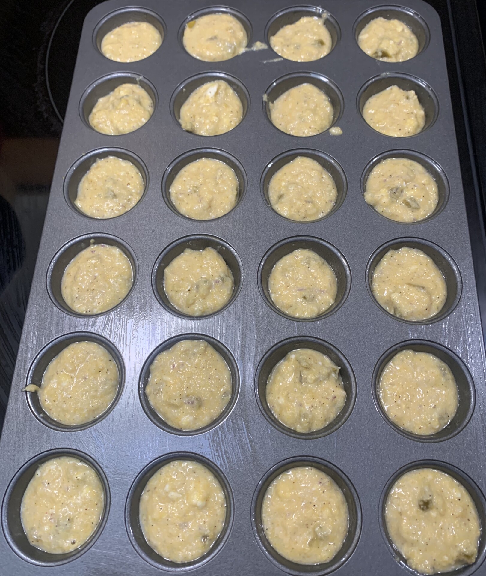 A mini muffin pan filled with raw muffin batter, ready to be baked. The batter appears to have small chunks, possibly fruit or nuts, and fills each cup almost to the top.