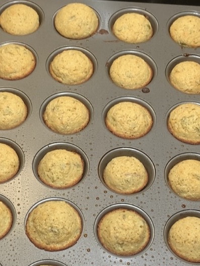 A muffin tin filled with freshly baked, golden brown muffins. The muffins are evenly risen and fill each cup of the tin, with some baking spray or oil visible on the pans surface.