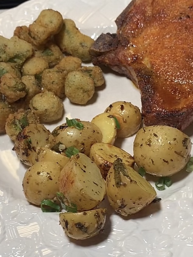A plate with roasted baby potatoes garnished with herbs, fried okra pieces, and a seasoned pork chop, all served on a white textured dish.