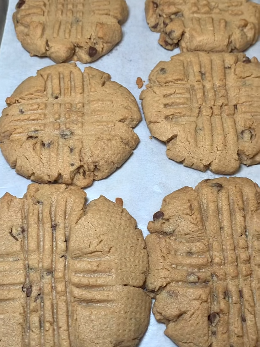 Six large peanut butter cookies with fork marks on top rest on a baking sheet. The cookies are slightly golden brown and have a rustic, homemade appearance.