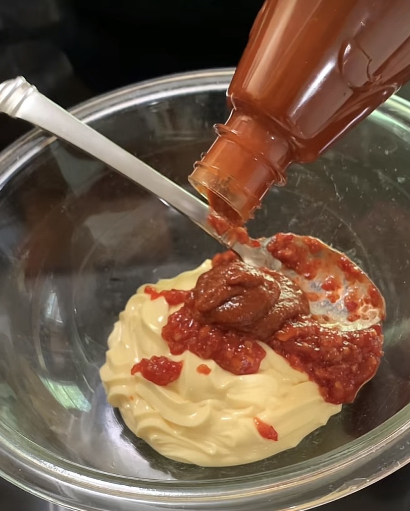 A close-up of a bowl containing a swirl of mayonnaise, with ketchup and chili sauce being squeezed on top. A spoon rests inside the bowl, ready for mixing.