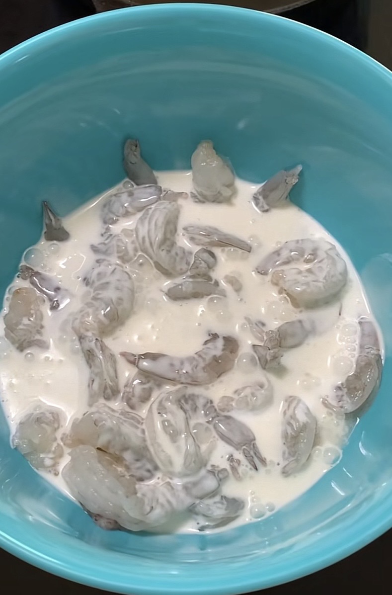 Raw shrimp soaking in a white liquid, likely milk or buttermilk, inside a blue bowl.
