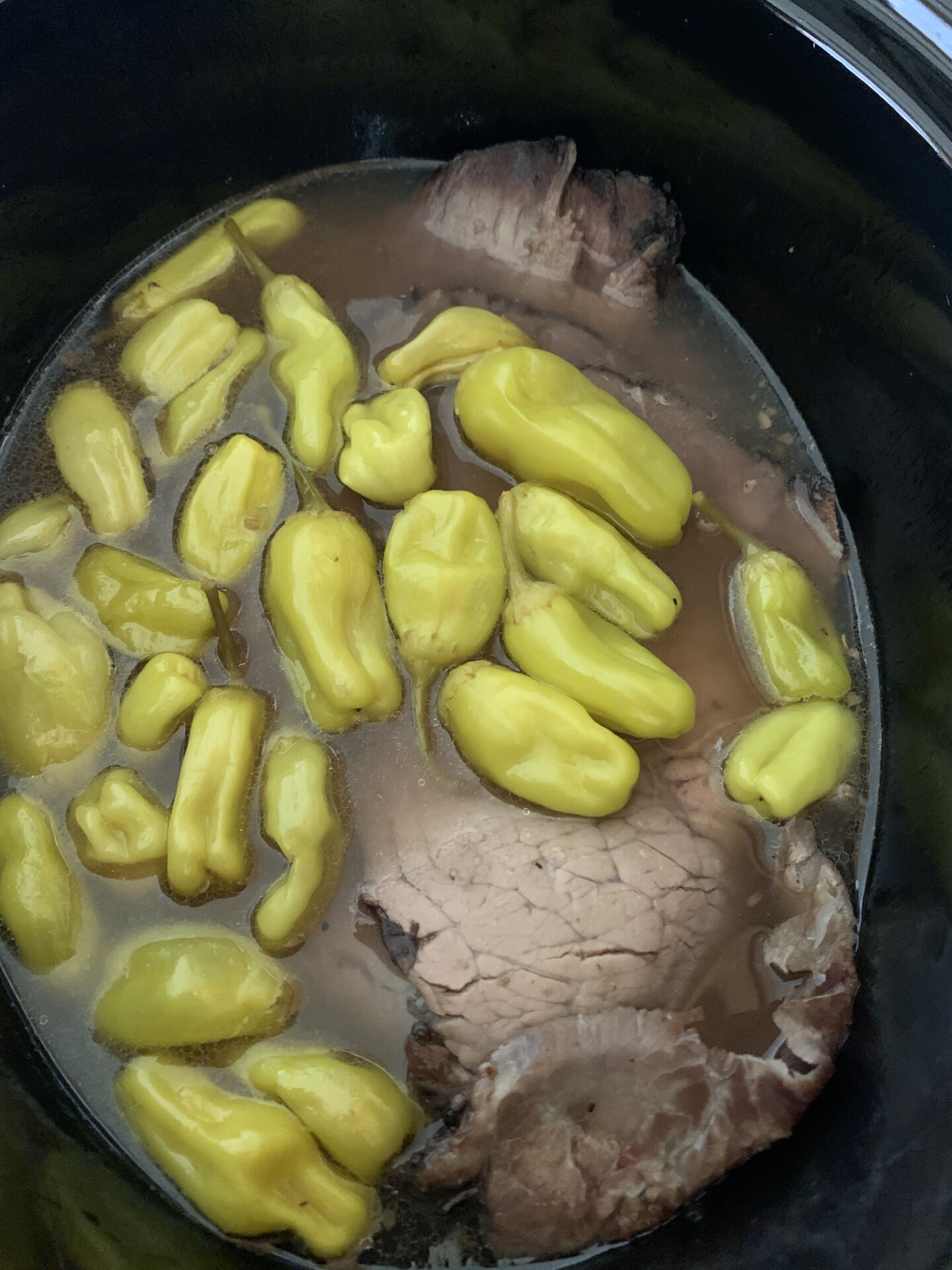 A beef roast cooking in a slow cooker, surrounded by whole yellow pepperoncini peppers and broth.