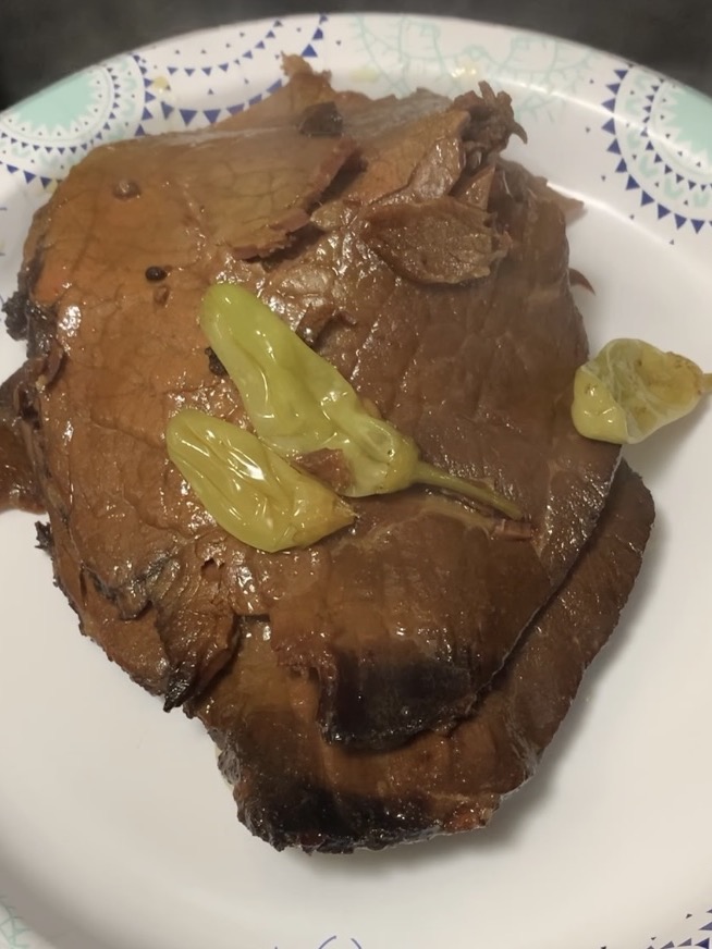Slices of cooked roast beef topped with several yellow pepperoncini peppers, served on a white paper plate with a blue and teal patterned rim.