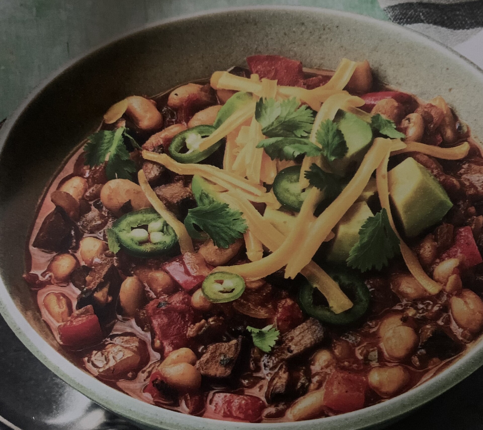 A bowl of chili topped with shredded cheese, sliced jalapeños, cilantro, and avocado cubes. The chili contains beans, tomatoes, and chunks of vegetables, all in a rich, red sauce.