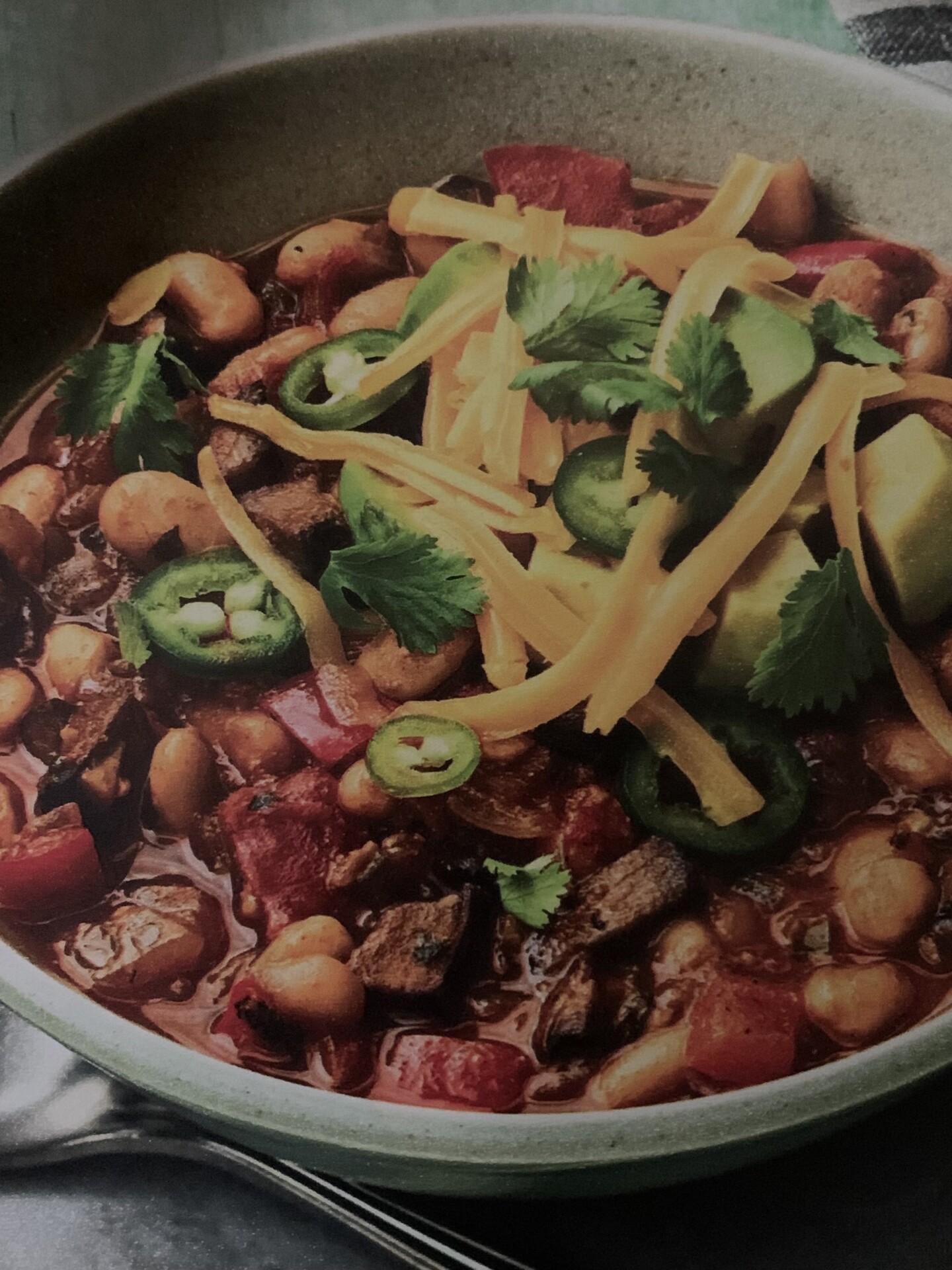 A bowl of chili topped with sliced jalapeños, shredded cheddar cheese, fresh cilantro, and chunks of avocado, featuring beans, tomatoes, and pieces of meat in a rich, hearty sauce.