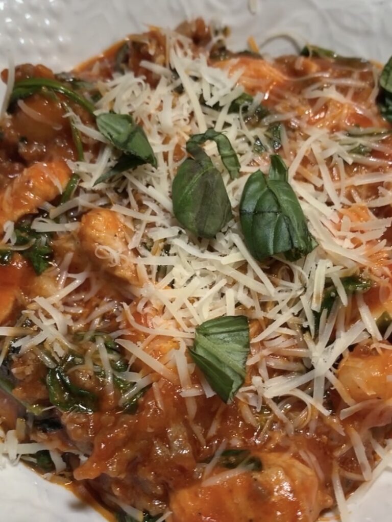 A close-up of pasta topped with tomato sauce, grilled chicken pieces, shredded parmesan cheese, fresh basil leaves, and spinach on a white plate.