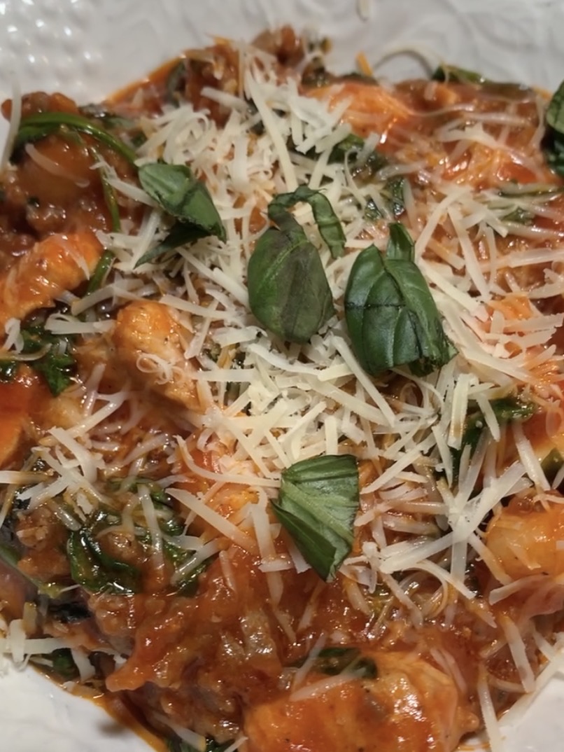 A close-up of pasta topped with tomato sauce, grilled chicken pieces, shredded parmesan cheese, fresh basil leaves, and spinach on a white plate.