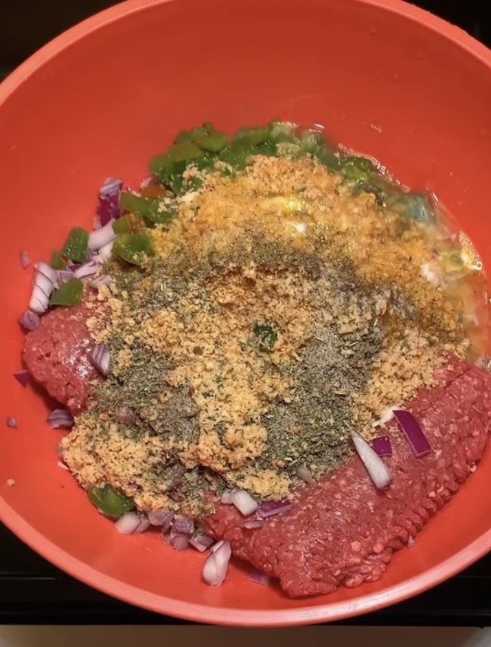 A red bowl contains raw ground beef, chopped green peppers, diced red onions, breadcrumbs, dried herbs, and seasonings, ready to be mixed for a recipe such as meatloaf or meatballs.