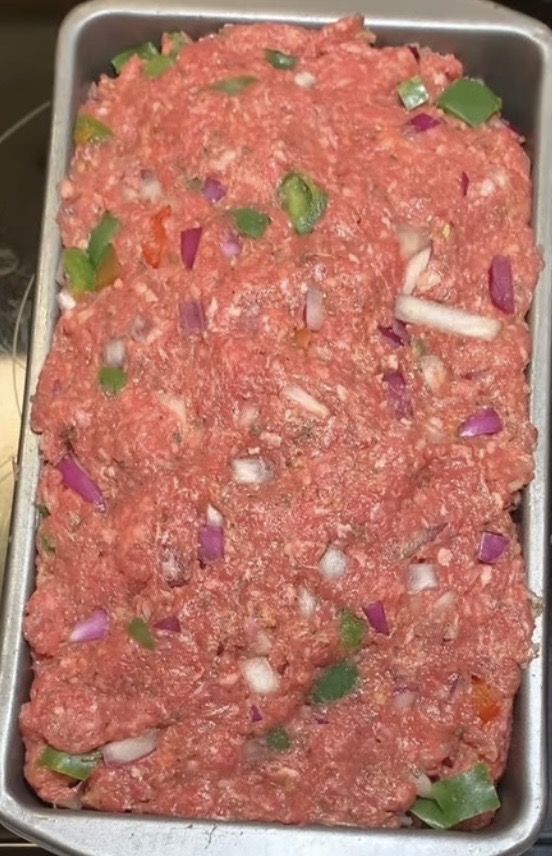 A metal loaf pan filled with raw meatloaf mixture containing ground meat, chopped red onions, green bell peppers, and other vegetables.
