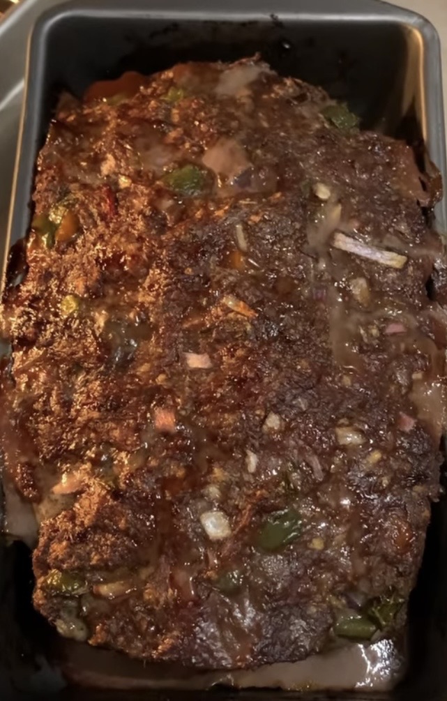 A close-up of a cooked meatloaf in a rectangular baking pan, showing a browned and textured surface with visible chunks of vegetables mixed throughout.