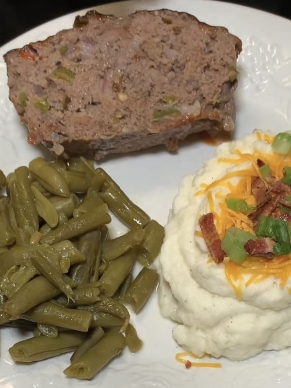 A white plate with a serving of meatloaf, mashed potatoes topped with shredded cheese, bacon bits, and green onions, and a side of cooked green beans.