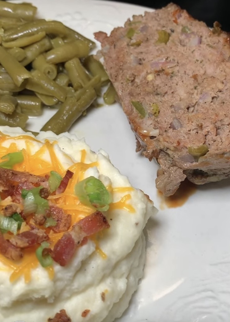 A plate of food with green beans, a slice of meatloaf, and mashed potatoes topped with shredded cheese, bacon bits, and chopped green onions.