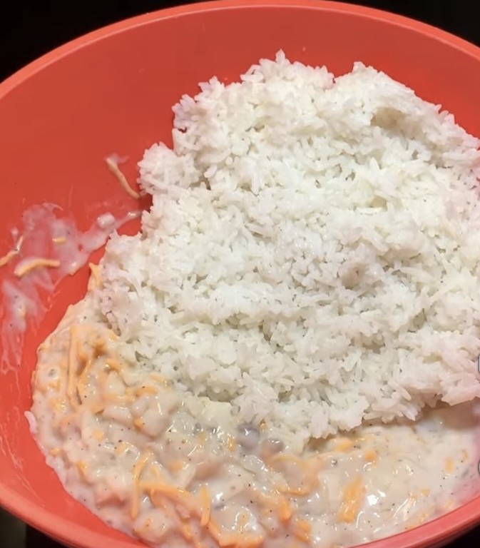 A red mixing bowl containing cooked white rice on top of a creamy mixture with shredded cheese.