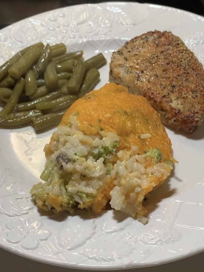 A white plate with green beans, a breaded pork chop, and a cheesy rice casserole with vegetables, including visible broccoli and mushrooms.