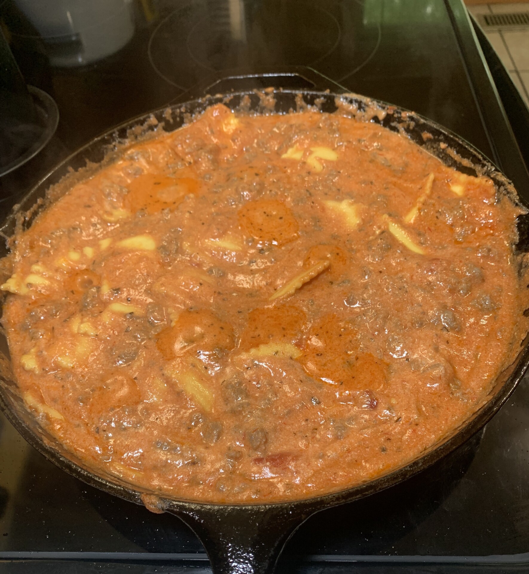 A cast iron skillet filled with a creamy, orange-red tomato-based sauce containing chunks of cheese and possibly mushrooms, simmering on a stovetop.