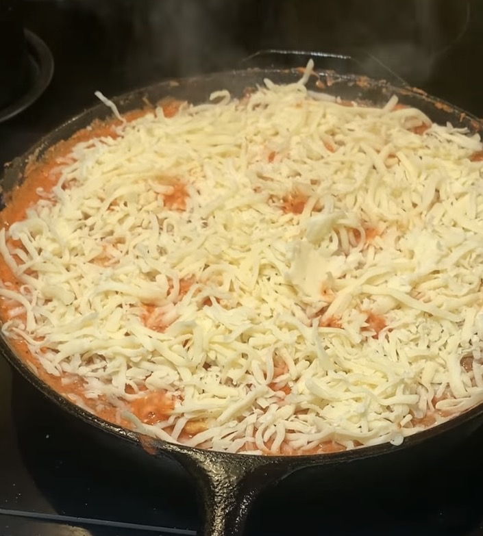 A cast iron skillet filled with a tomato-based dish topped with a generous layer of shredded cheese, starting to melt from the heat. Steam rises from the hot food, indicating it is being cooked.