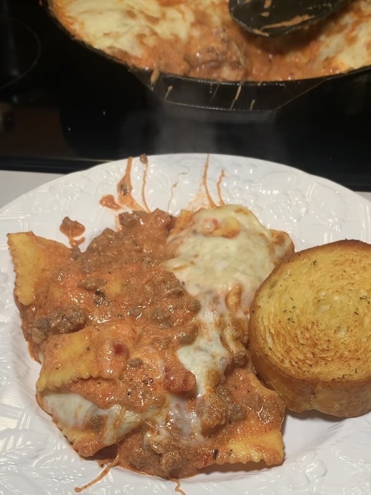 A plate with cheesy baked pasta in tomato meat sauce, next to a slice of garlic bread. In the background, a pan of the same pasta dish with melted cheese on top is visible. Steam is rising from the food.