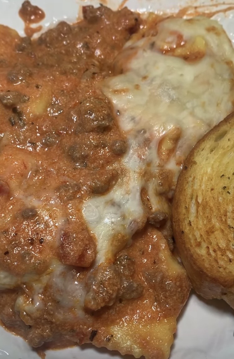 Close-up of cheesy baked lasagna with a meat and tomato sauce, next to a slice of toasted garlic bread on a white plate.