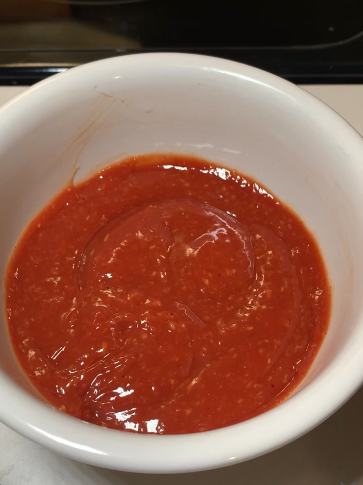 A white bowl filled with a smooth, bright red sauce sits on a countertop. The sauce appears thick and glossy, with a slightly textured surface.