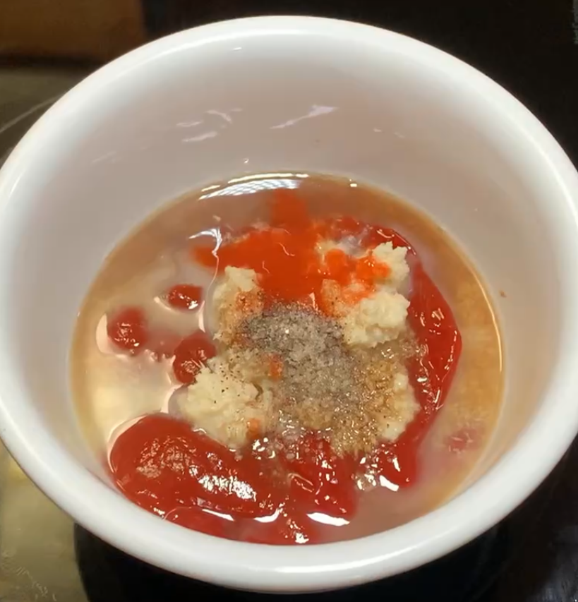 A white bowl containing ketchup, horseradish, hot sauce, ground black pepper, and a light brown liquid, likely being prepared for a sauce or dip.