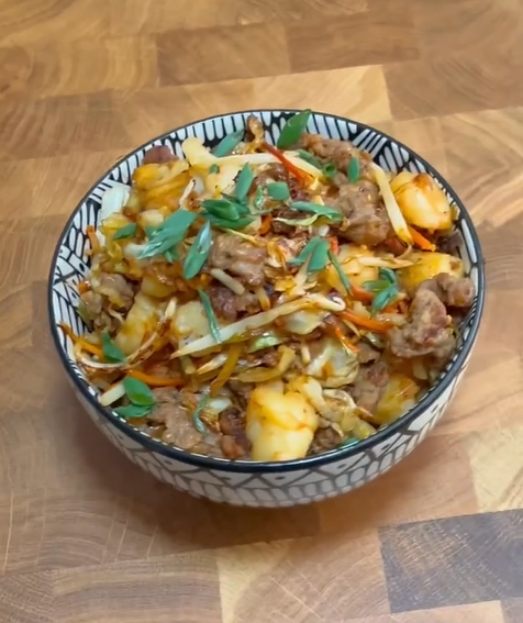 A patterned bowl filled with a stir-fry of meat, cabbage, carrots, and potatoes, garnished with fresh herbs, sits on a wooden surface.