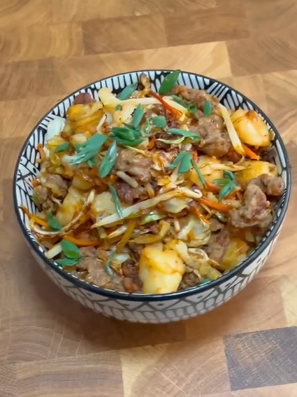 A patterned bowl filled with a hearty dish of diced potatoes, beef, shredded carrots, and onions, garnished with fresh green herbs, sits on a wooden surface.