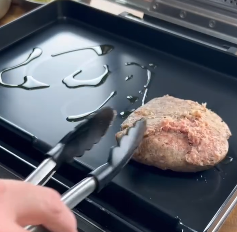 A hand holds metal tongs, placing a raw ground meat patty onto a lightly oiled flat grill surface. The grill has drizzled oil on it, ready for cooking.