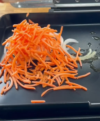 A pile of julienned carrots and sliced onions on a black griddle with a small amount of oil, ready to be cooked.