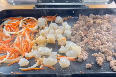 Vegetables, shrimp, and ground meat cook side by side on a flat-top grill, with steam rising from the sizzling food.