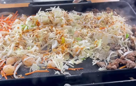 A sizzling grill topped with shredded cabbage, carrots, shrimp, ground meat, and sauce, creating a colorful and steamy stir-fry in progress.