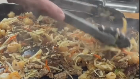 A close-up of a sizzling stir-fry with beef, shrimp, cabbage, and carrots being mixed with tongs on a hot grill, with steam rising from the food.
