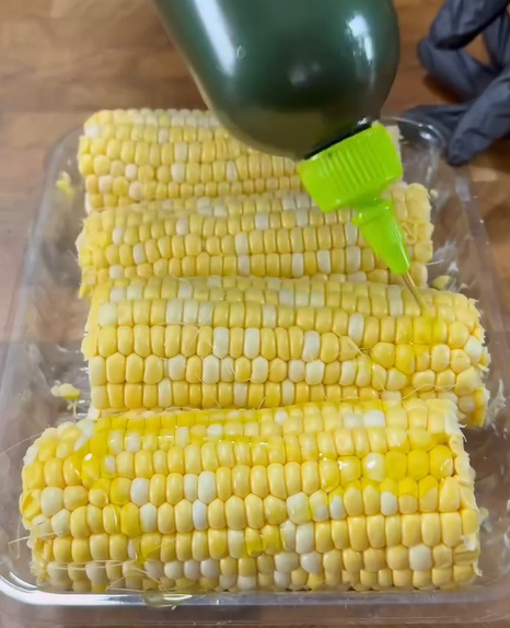 Four ears of corn on the cob in a clear dish are being drizzled with oil from a green bottle. The corn rests on a wooden surface, and a gloved hand is partially visible in the background.