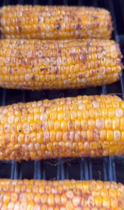 Close-up of several ears of corn on the cob being grilled, showing a golden-yellow color with char marks and seasoning on the surface.