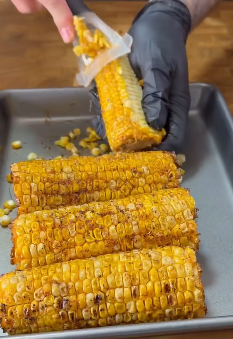 A hand in a black glove uses a grater to shave kernels off a seasoned, cooked corn cob over a tray holding three other spiced corn cobs on a wooden surface.